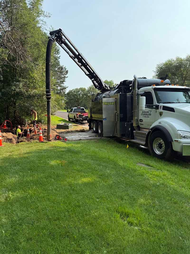 Truck with extended arm vacuuming ground near a hole, in a grassy area with trees, under a bright sky.
