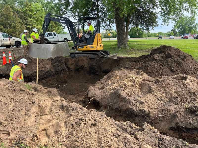 A yellow excavator lifts a concrete structure over a deep dirt excavation site while workers in safety gear oversee work.