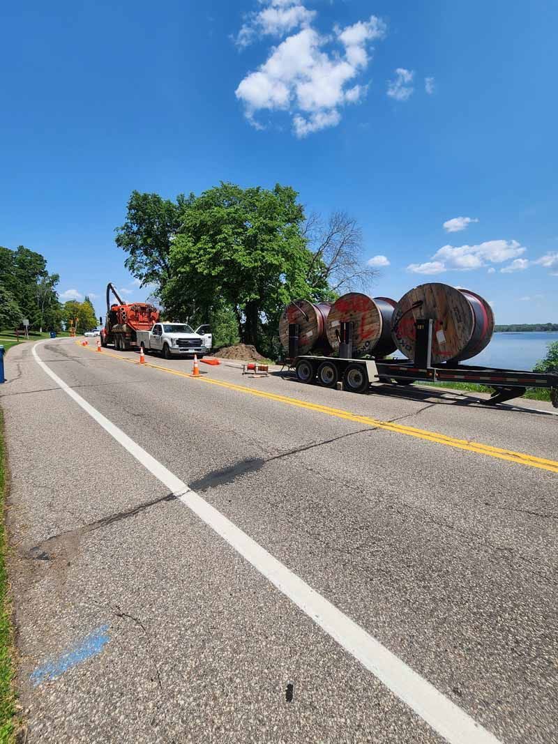 Roadside scene: truck with large spools of cable, work vehicles, traffic cones, and water in the background. Sunny day.