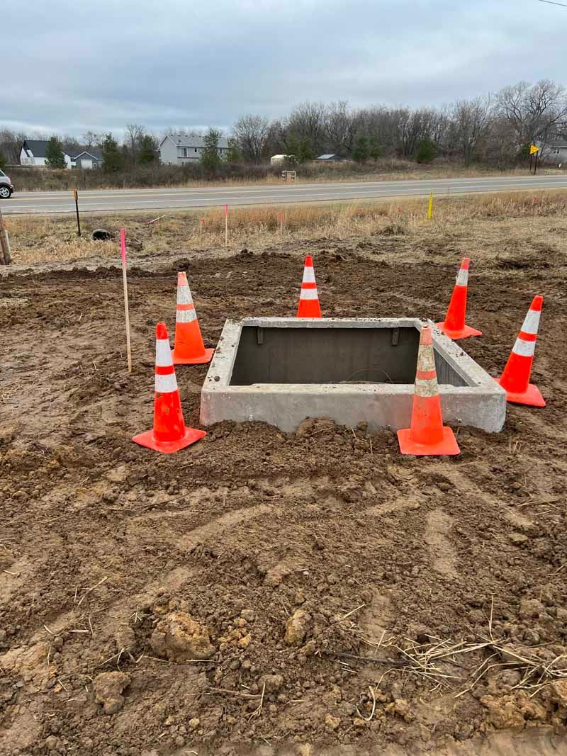 An open concrete utility access point surrounded by orange safety cones on a dirt construction site.
