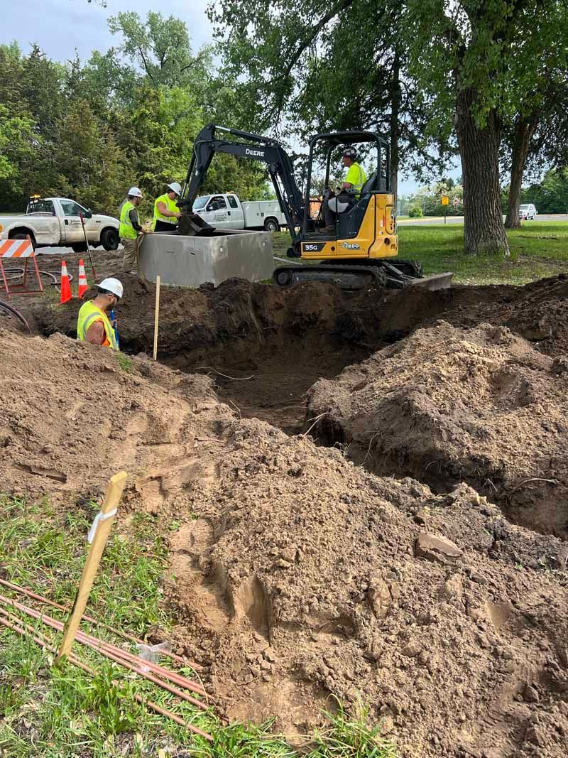 Construction workers and a backhoe excavate near a concrete ring in a dirt pit outdoors.