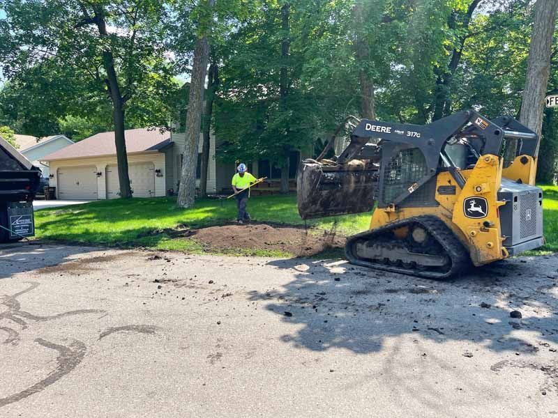 Yellow skid steer dumping dirt near a worker in front of houses with green lawns.