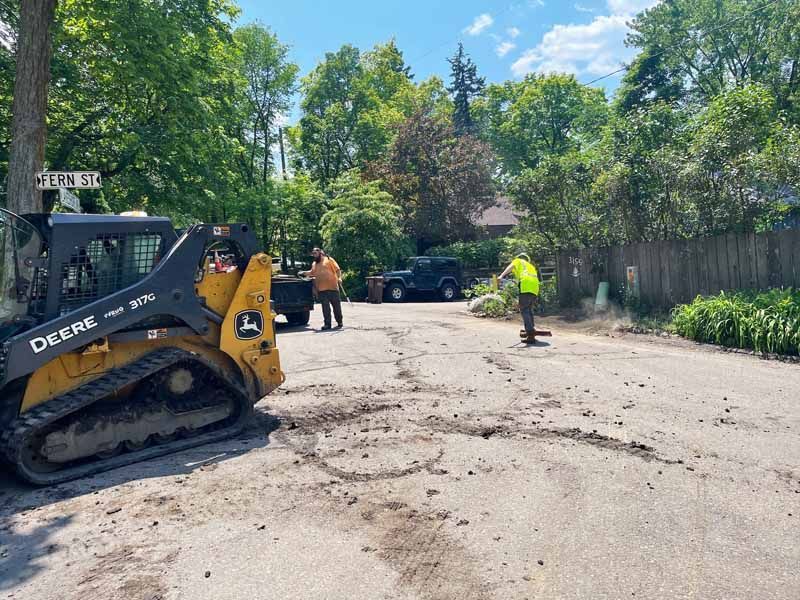Yellow John Deere skid steer; workers on asphalt road with debris. Trees in background, sunny day.