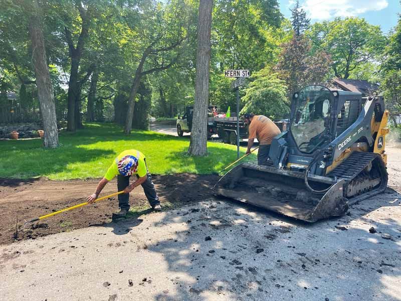 Construction workers and skid steer machine on a residential street, digging dirt in sunny weather.