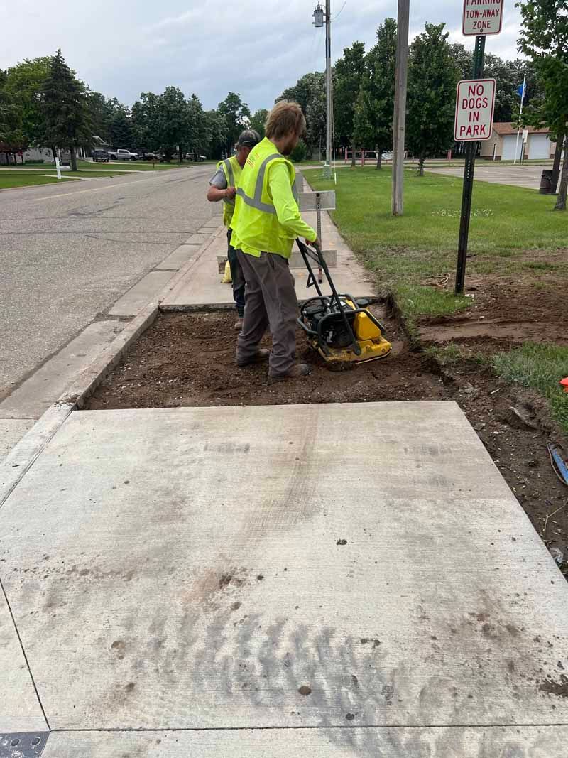 Workers in reflective vests compact dirt with a machine, preparing for sidewalk repair.