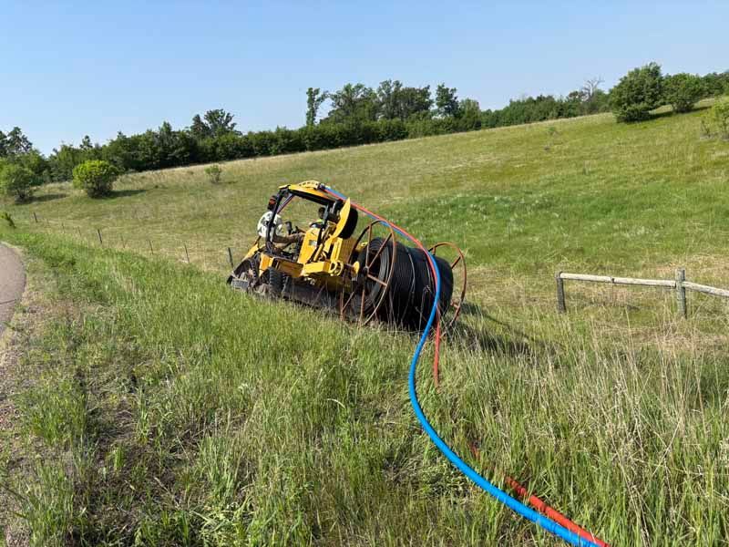 Yellow construction machine off-road, laying blue and red cable in a grassy field on a sunny day.