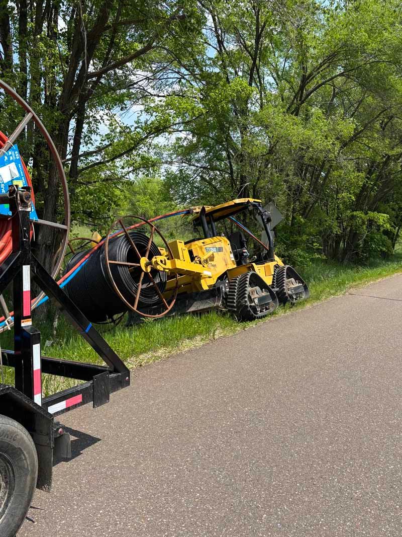 Yellow construction vehicle on tracks next to a large black cable reel on a trailer beside a paved road.