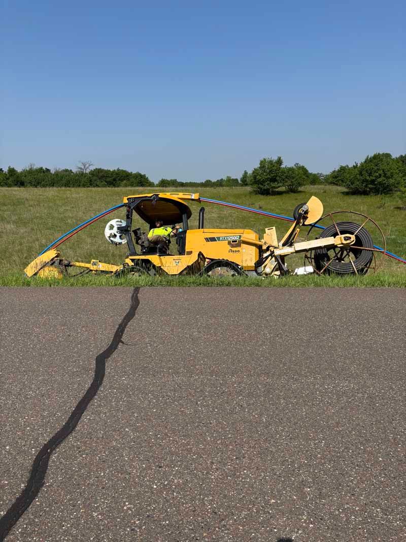 Yellow mowing machine with operator, cutting grass next to a paved road on a sunny day.