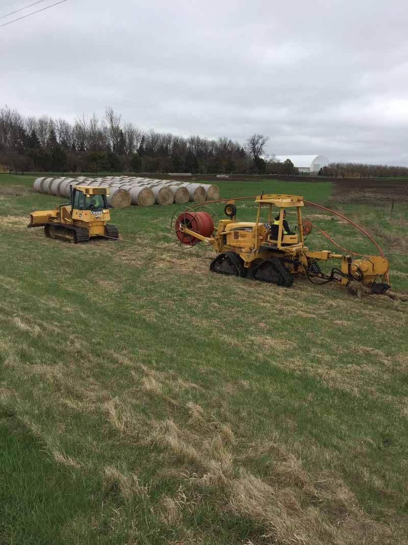 Two yellow construction vehicles in a field, with hay bales in the background under a cloudy sky.