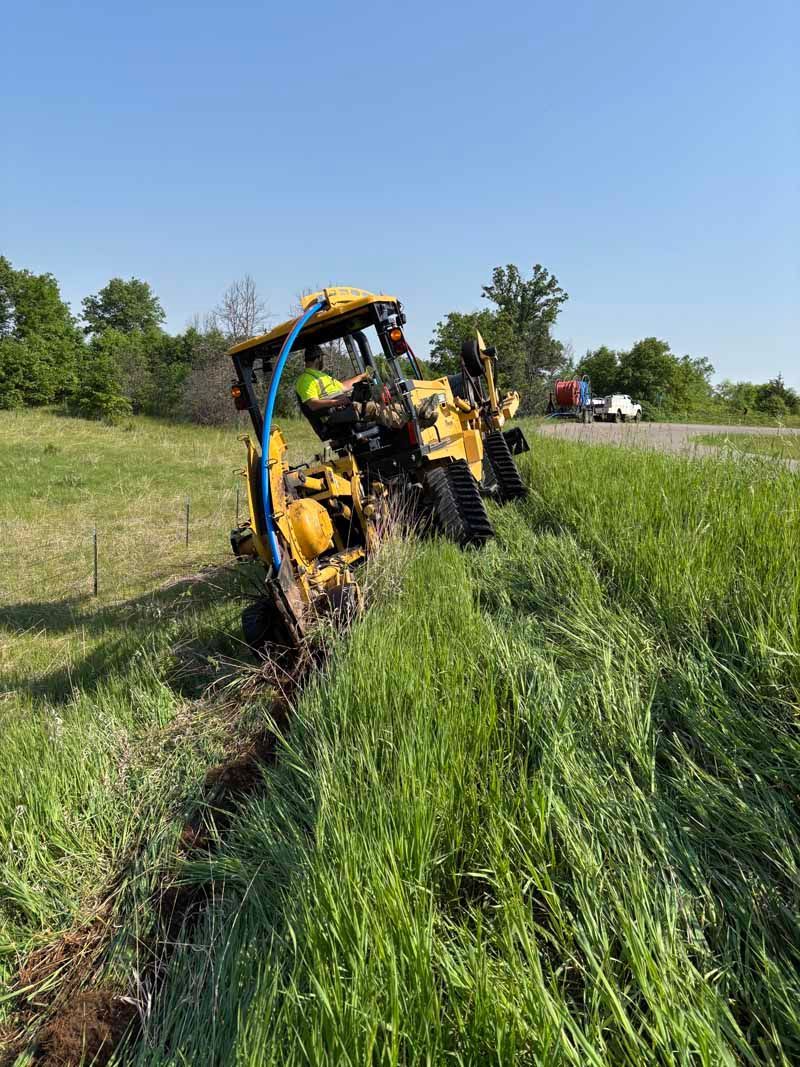 Yellow construction vehicle on tracks cutting a trench in tall grass beside a road, blue sky.