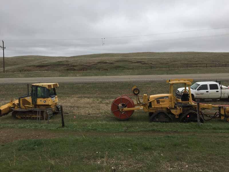 Two yellow tractors with a cable reel in a field beside a road and a white truck. Overcast sky.
