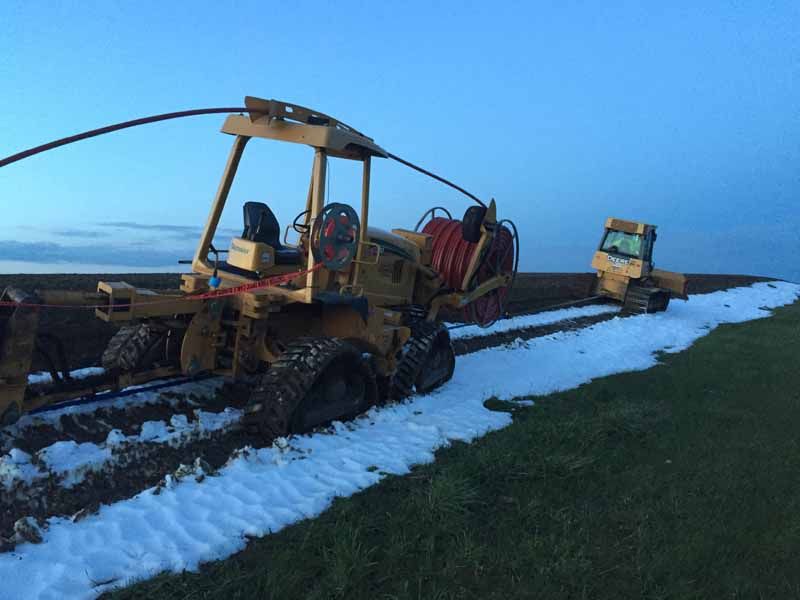 Two yellow trenching machines laying cable in a snowy field at dusk.