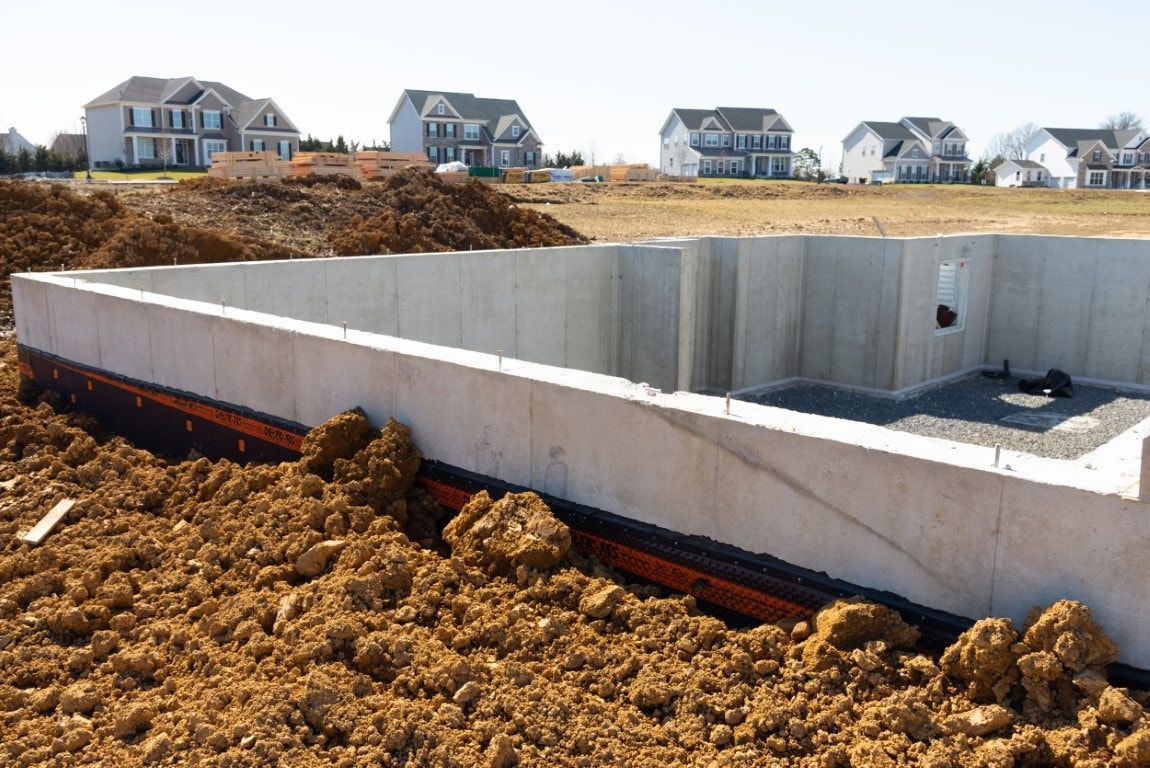 Concrete foundation of a house under construction with surrounding dirt and houses in the background.