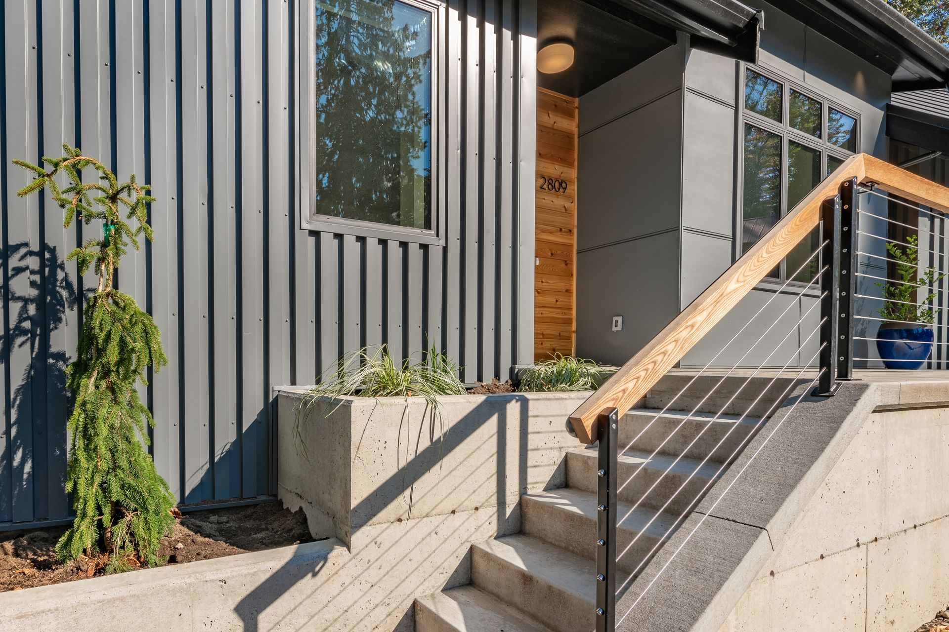 Modern house entryway with metal siding, concrete steps, and wooden handrail in sunlight