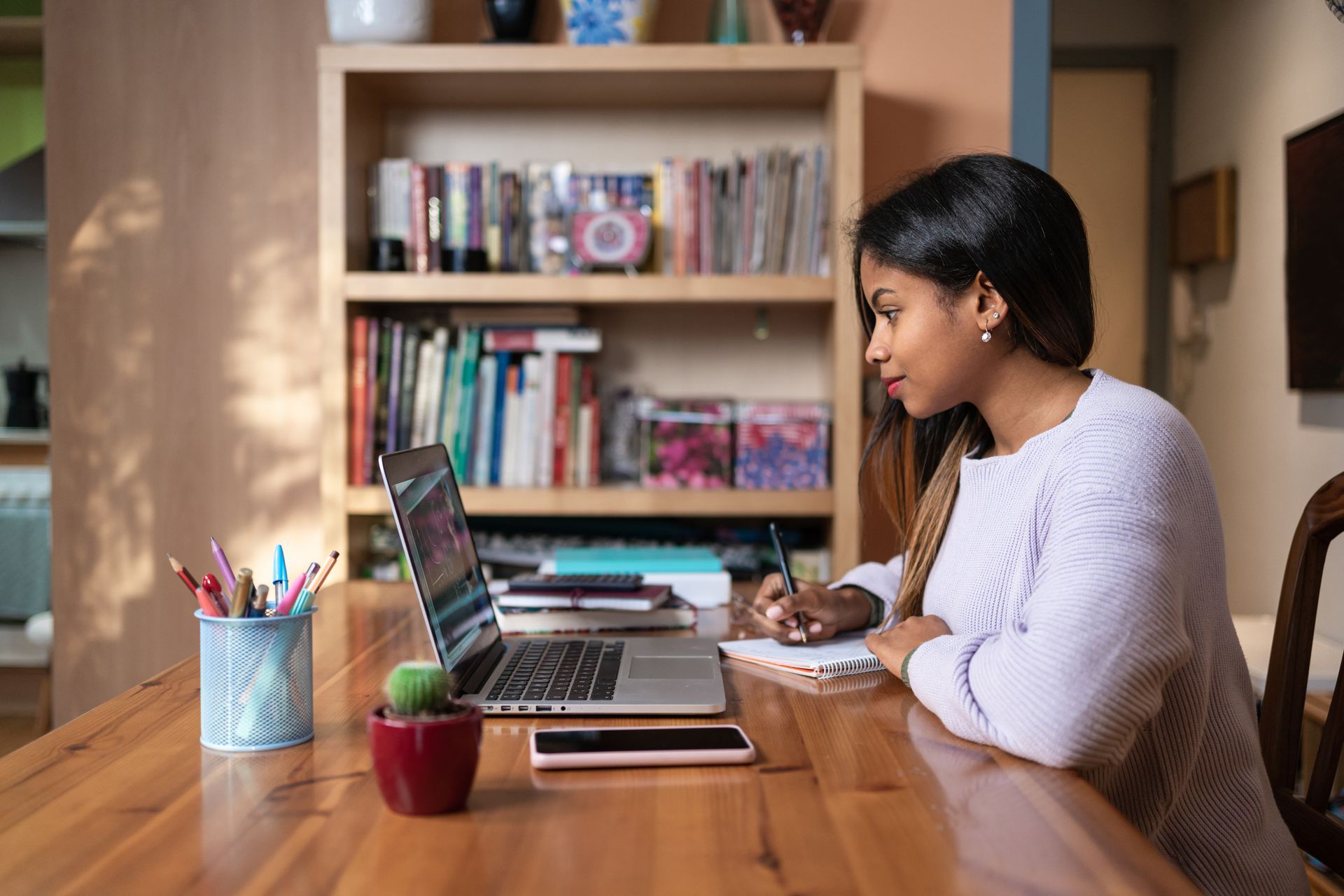 Woman working on laptop, taking notes at a wooden table in a home office. Woman working on laptop, taking notes at a wooden table in a home office.