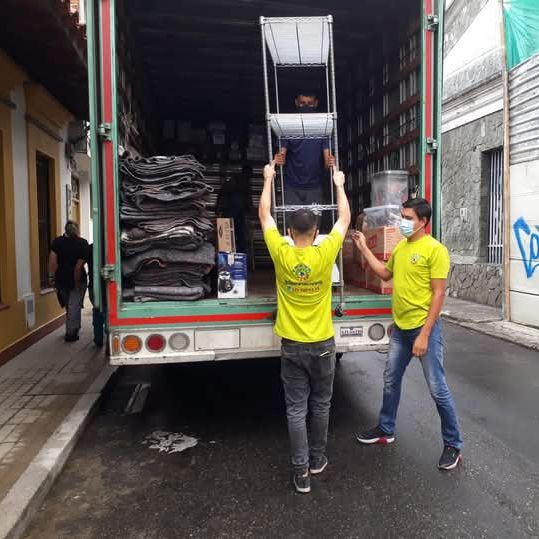 Dos trabajadores con camisas amarillas descargan sillas apiladas de un camión en un callejón estrecho.