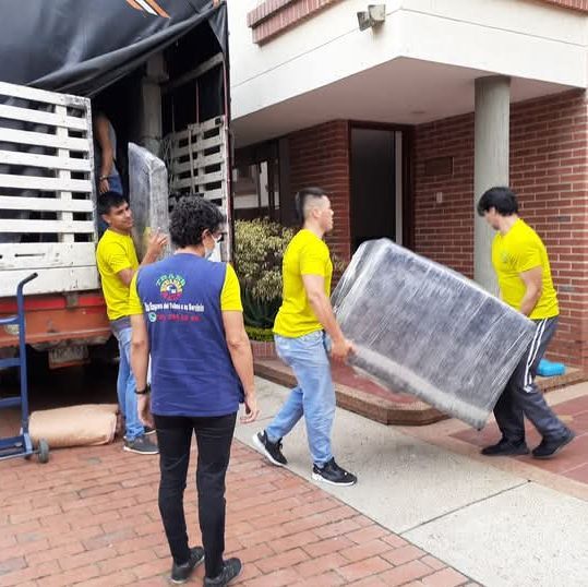Cuatro trabajadores trasladan un colchón envuelto sobre una carretilla cerca de la entrada de un edificio.