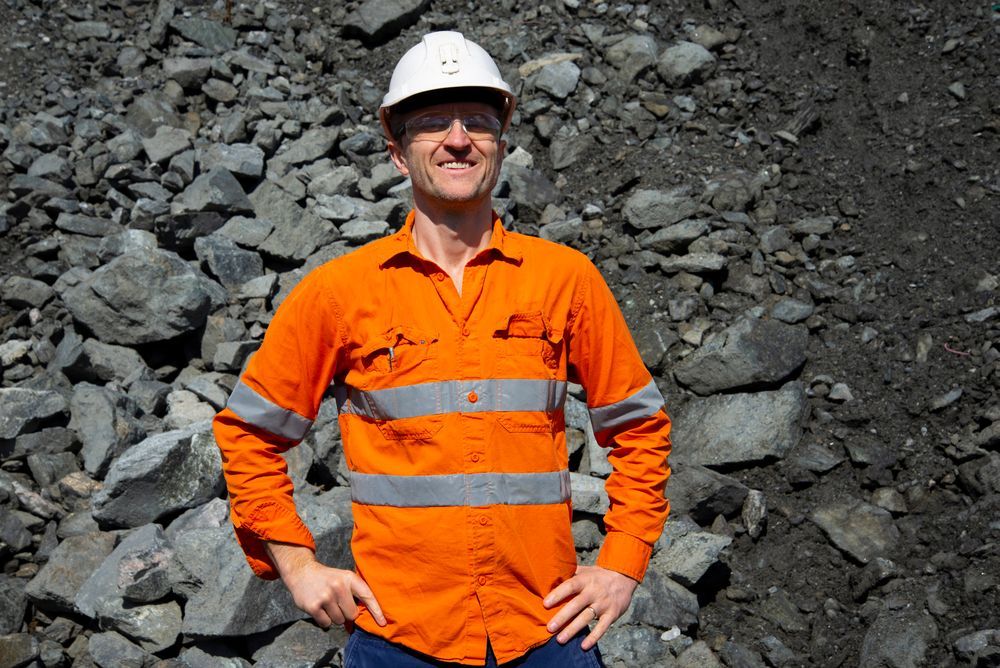A Man Wearing an Orange Shirt and a Hard Hat is Standing in Front of a Pile of Rocks — Calibre Resourcing Solutions in Cairns, QLD