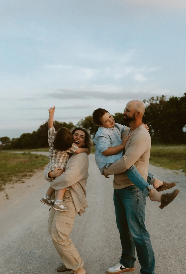 Family of three standing outdoors at sunset, smiling and lifting a child in the air near a grassy path.