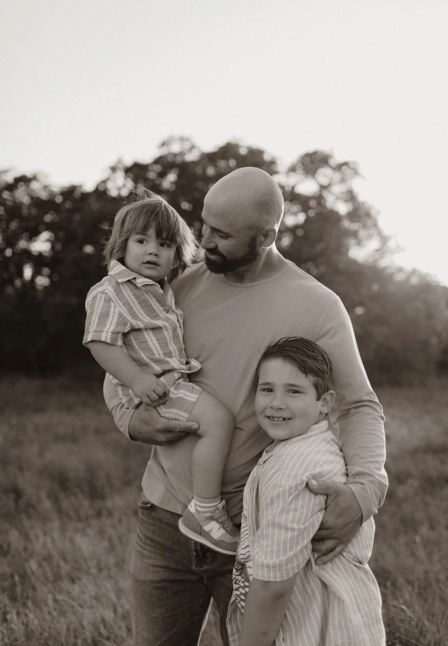 Man holding two children in a grassy field, with trees blurred in the background.