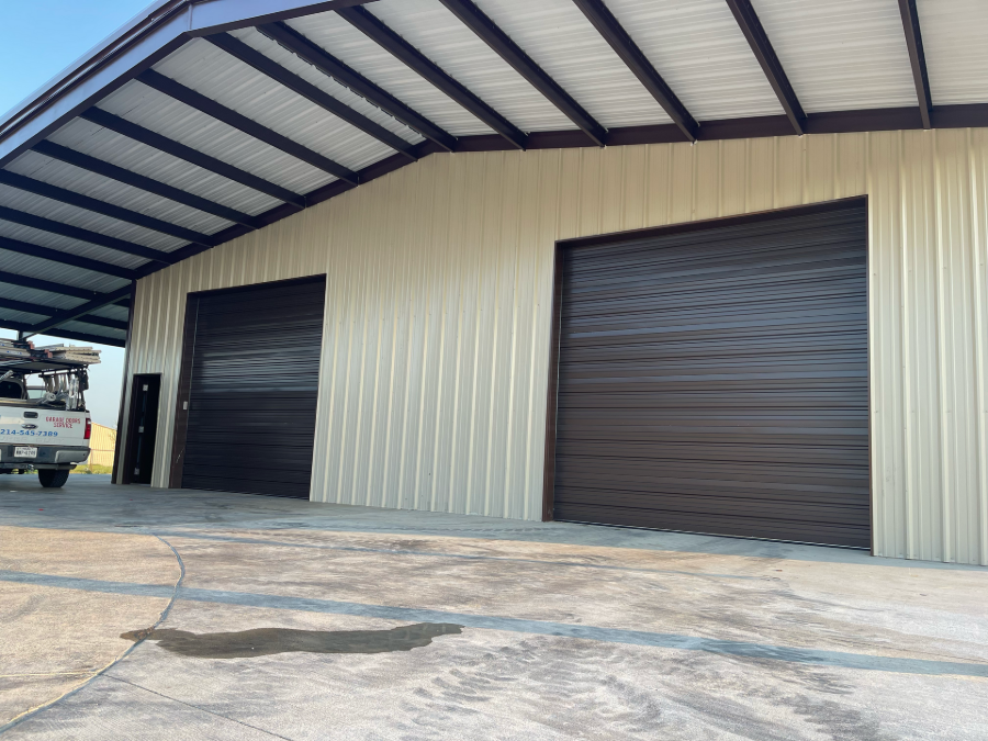 Warehouse exterior with beige metal siding, two dark roll-up doors, and a truck parked at left under a large roof overhang