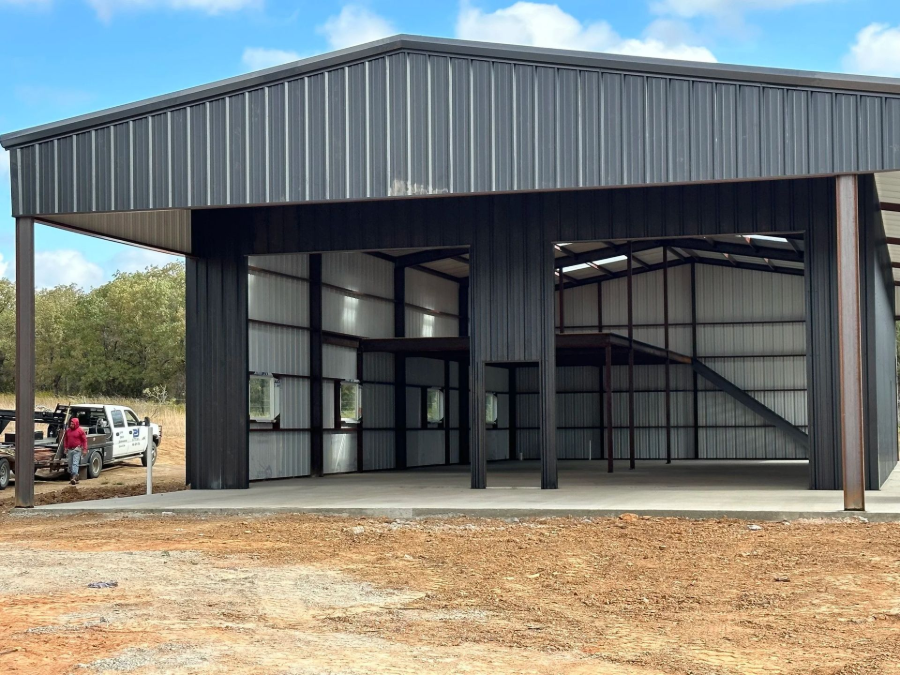 Open-sided metal pavilion with black beams and a corrugated roof on a gravel lot