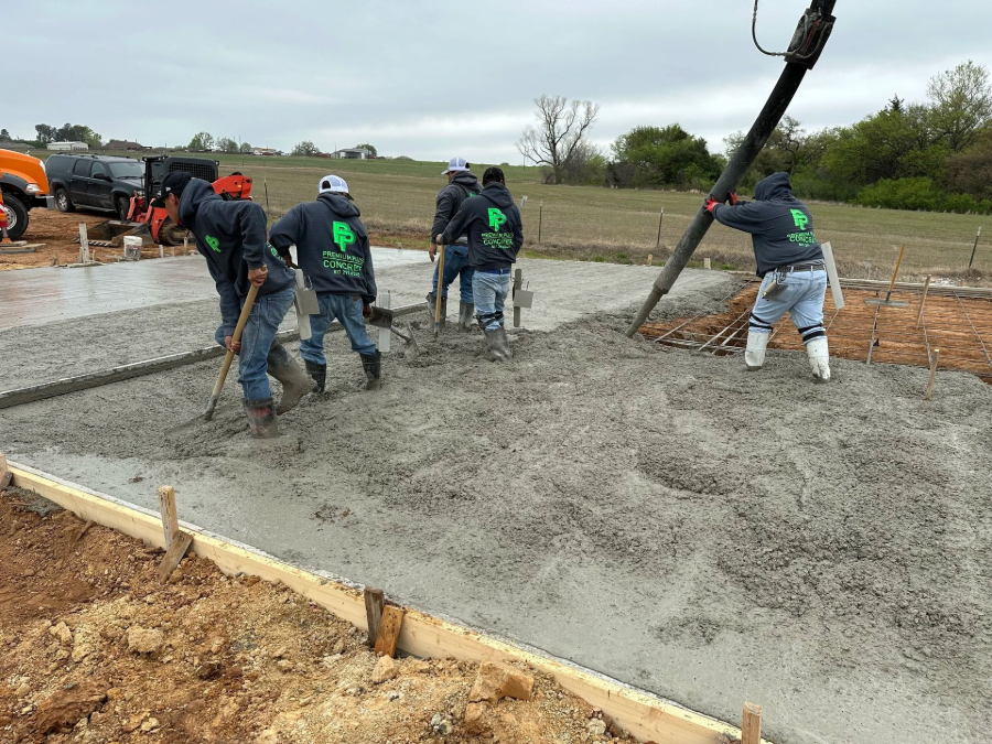 Workers pour and smooth concrete at a construction site, with a concrete pump hose and dirt forms nearby.
