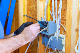 Electrician wiring a gray junction box in a wall stud bay with white cables and black gloves