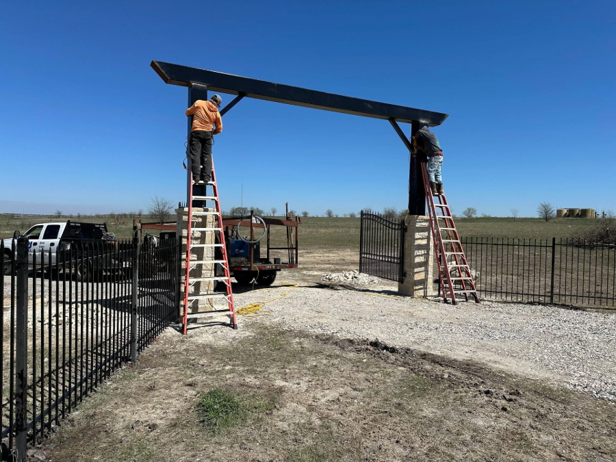 Two ladders support a beam at a fenced outdoor entrance under a clear blue sky