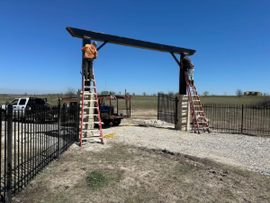 Workers standing on ladders installing a large metal gate frame at a fenced entrance under a blue sky