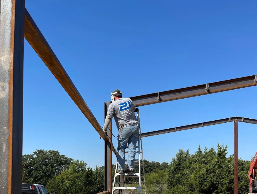 Person on a ladder painting a rusty metal frame against a clear blue sky.