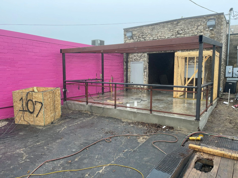 Outdoor patio with a black metal canopy beside a bright pink wall and a stone building, with hoses on the ground