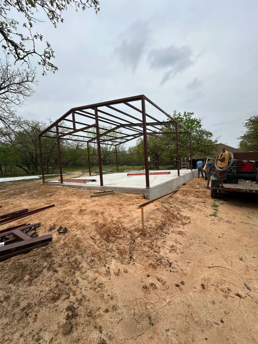 Steel frame building under construction on a concrete slab, with dirt foreground and a truck at right.