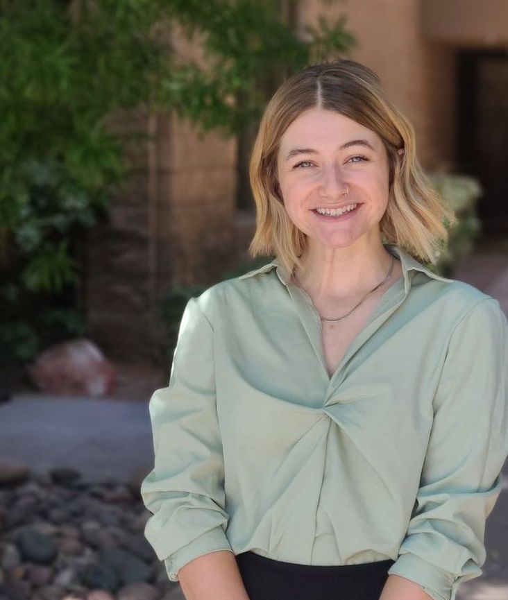 Smiling person in a light green blouse standing outdoors near plants and a brick wall.