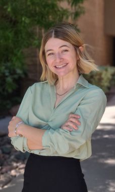 Smiling person in a light green shirt and black pants standing outdoors with arms crossed