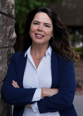 Woman standing outdoors by a tree, arms crossed, wearing a white blouse and navy cardigan, smiling.