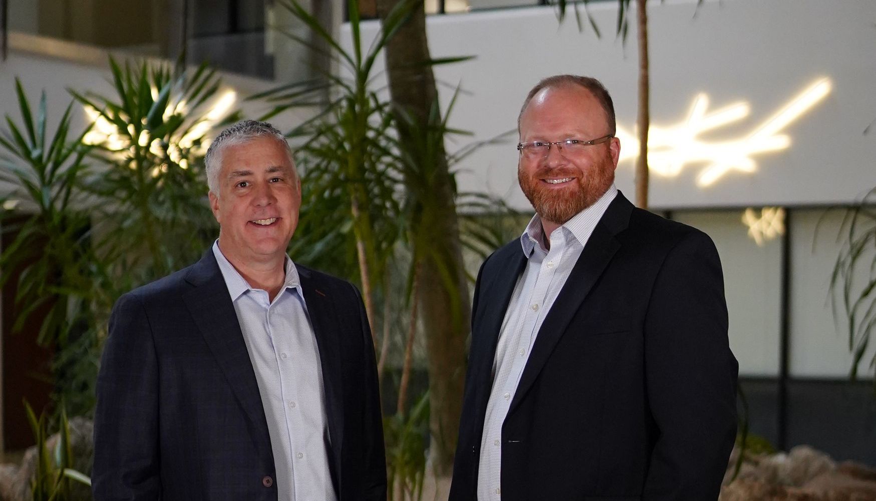 Two men in suits smiling indoors, with plants and a bright wall in the background.