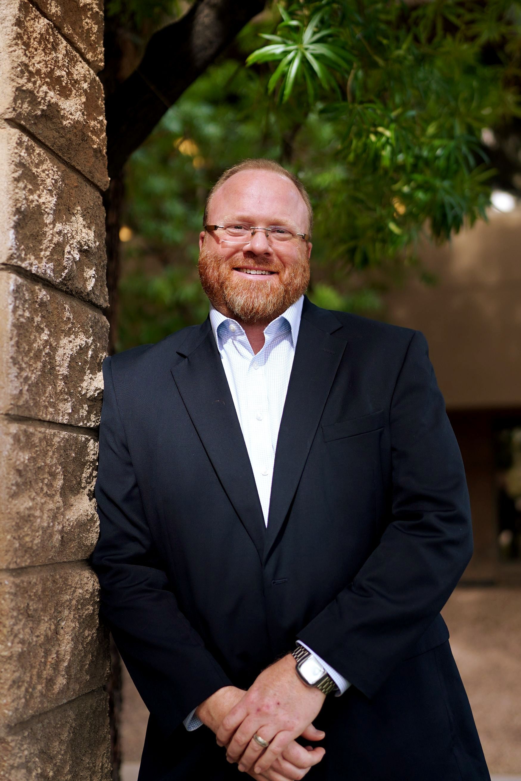 Smiling man in a dark suit standing outdoors beside a stone wall and greenery