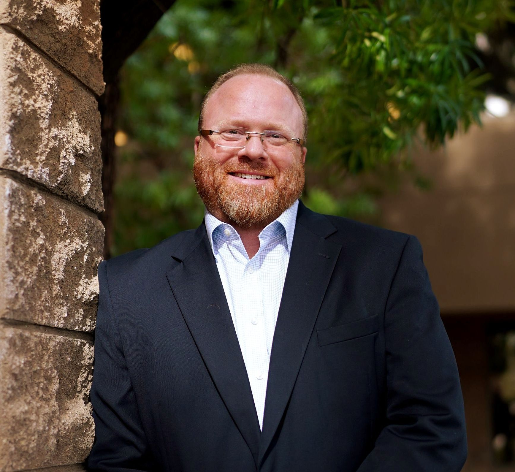 Smiling man in a dark suit standing outdoors beside a stone wall and greenery