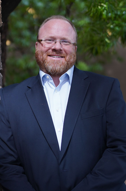 Smiling person in a dark suit standing outdoors with hands clasped.