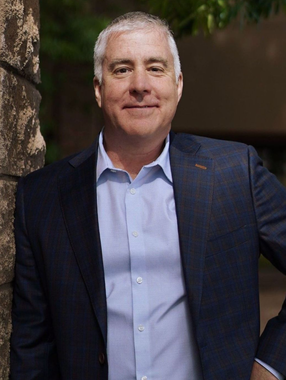 Man in a navy suit standing outdoors, smiling with one hand on his hip.