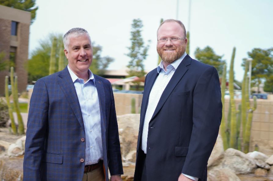 Two men in suits standing outdoors near rocks and trees, smiling at the camera.