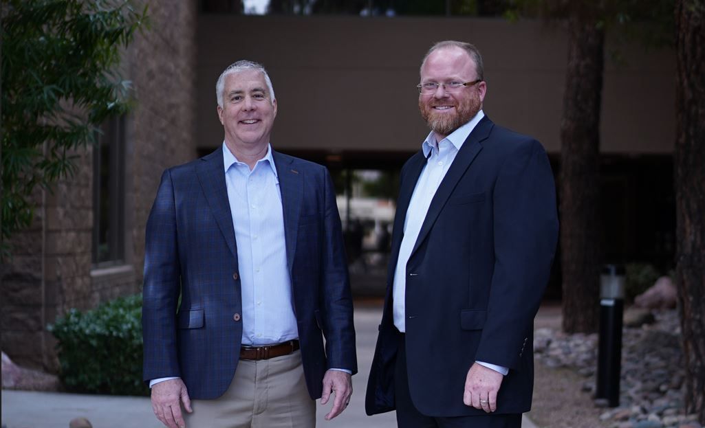 Two men in dark blazers standing and smiling outside a building entrance