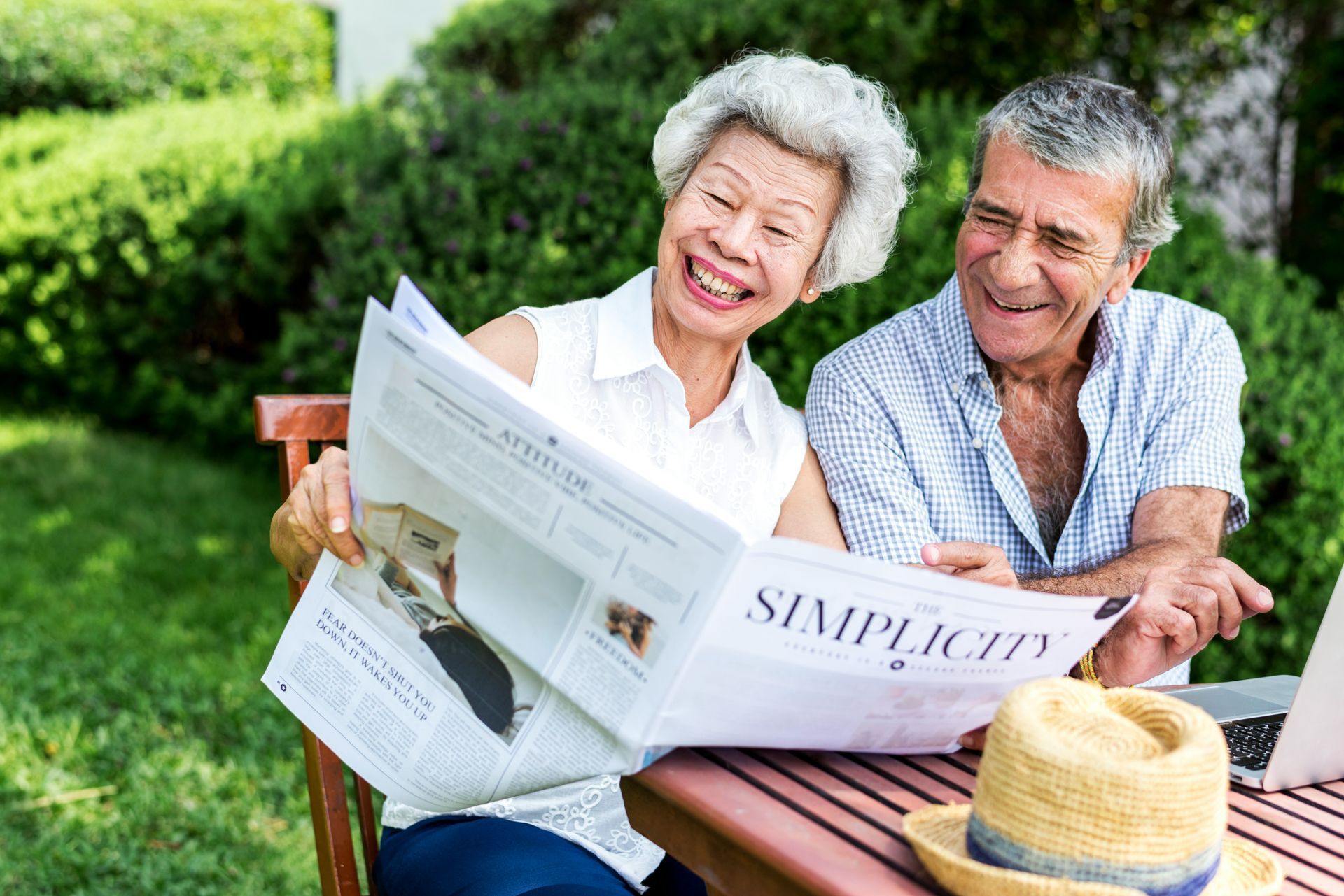 Smiling couple reading a newspaper at a park picnic table with a straw hat nearby