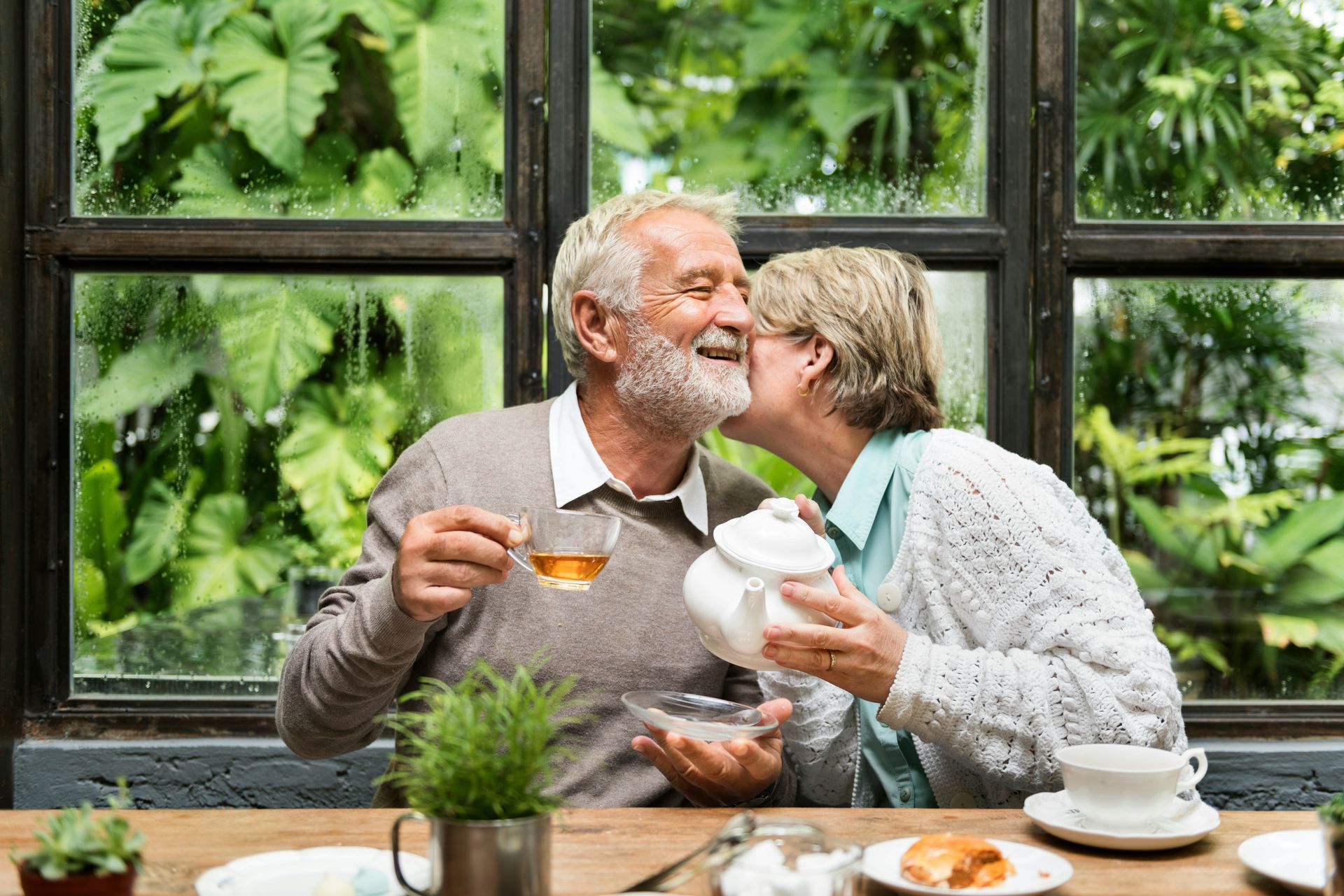 Two people sharing tea at a café table by a lush green window, smiling and kissing.
