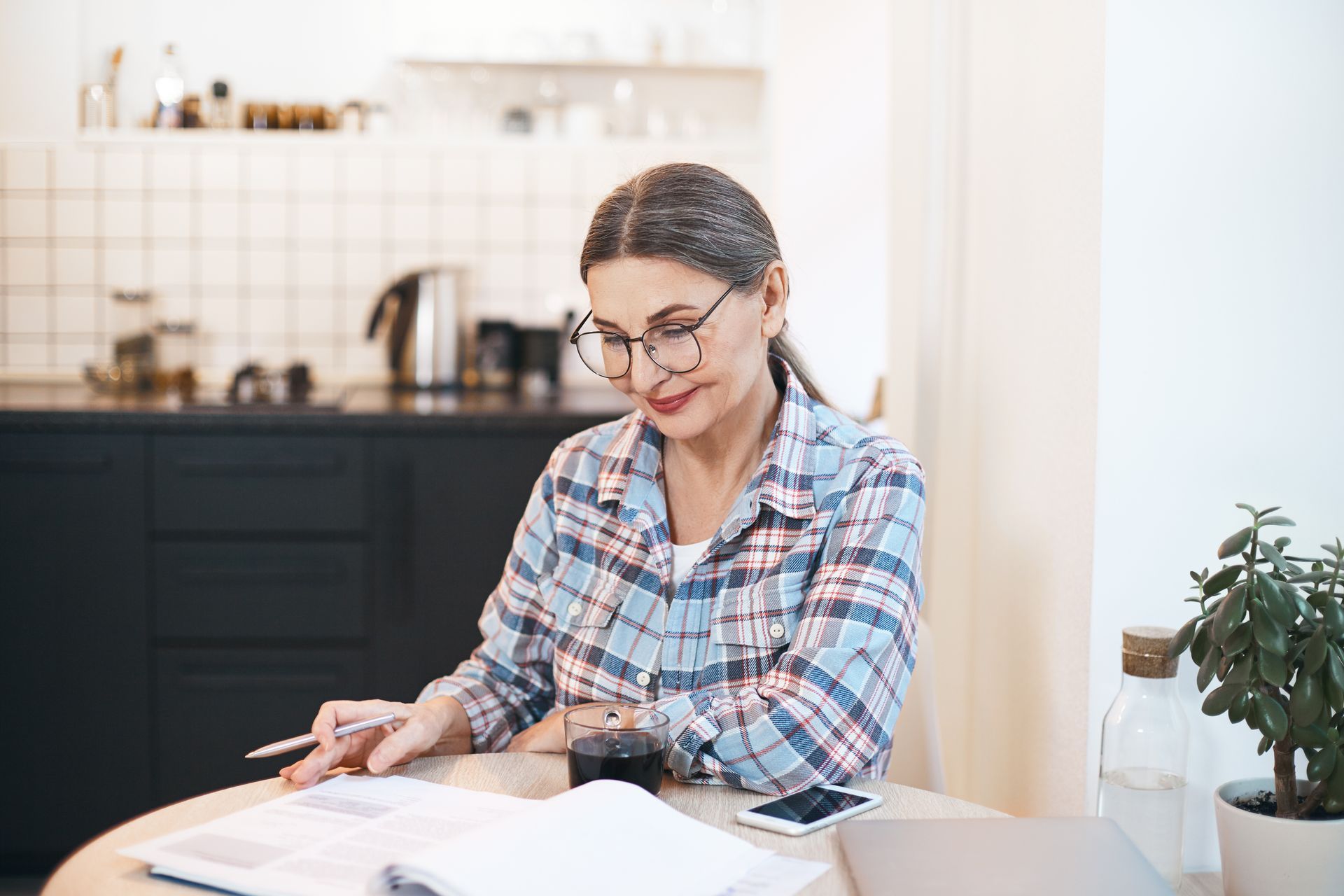 Person reading and writing at a table in a bright kitchen, wearing glasses and a plaid shirt.
