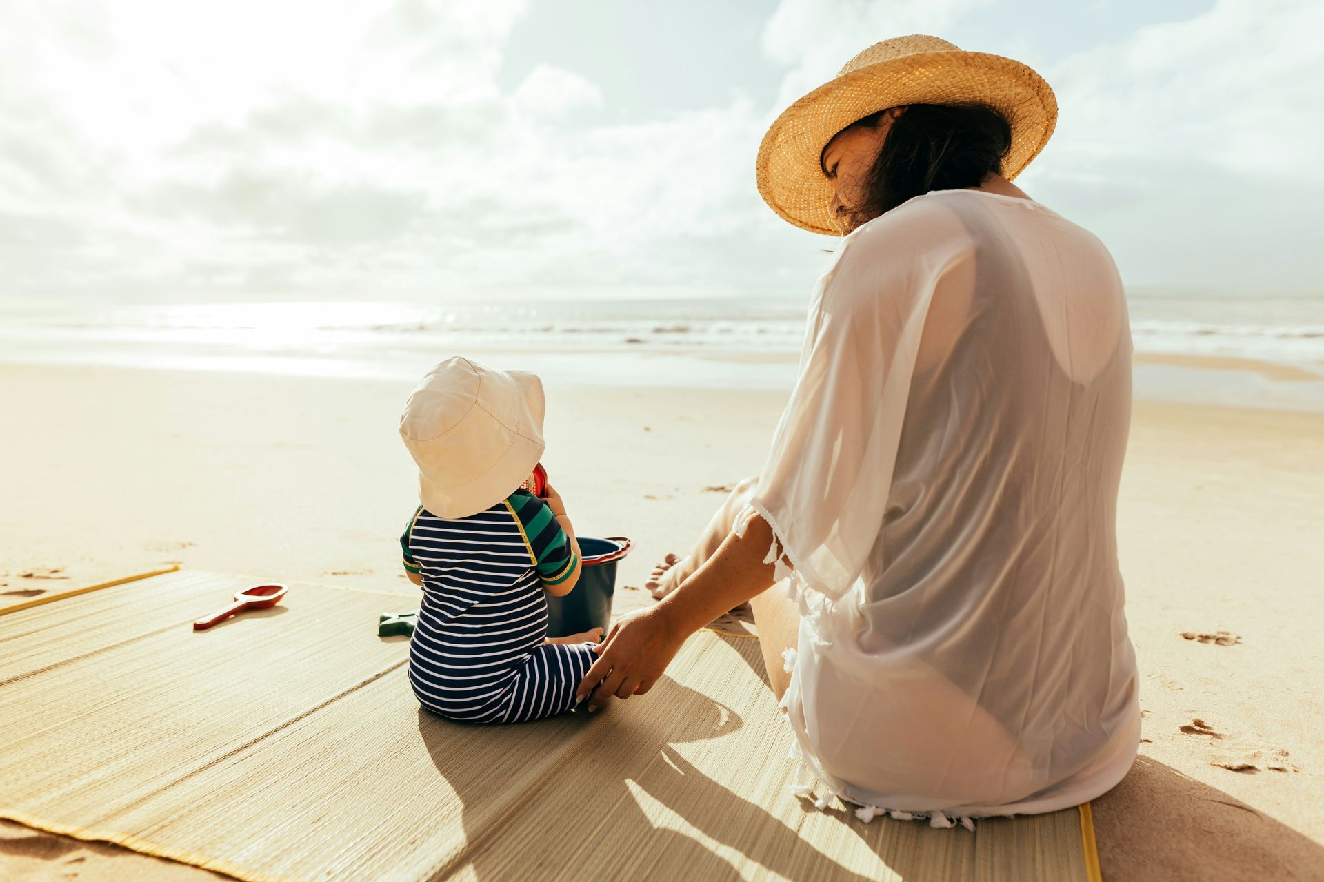 Two people sitting on a sunny beach, facing the ocean, wearing light clothes and a straw hat.
