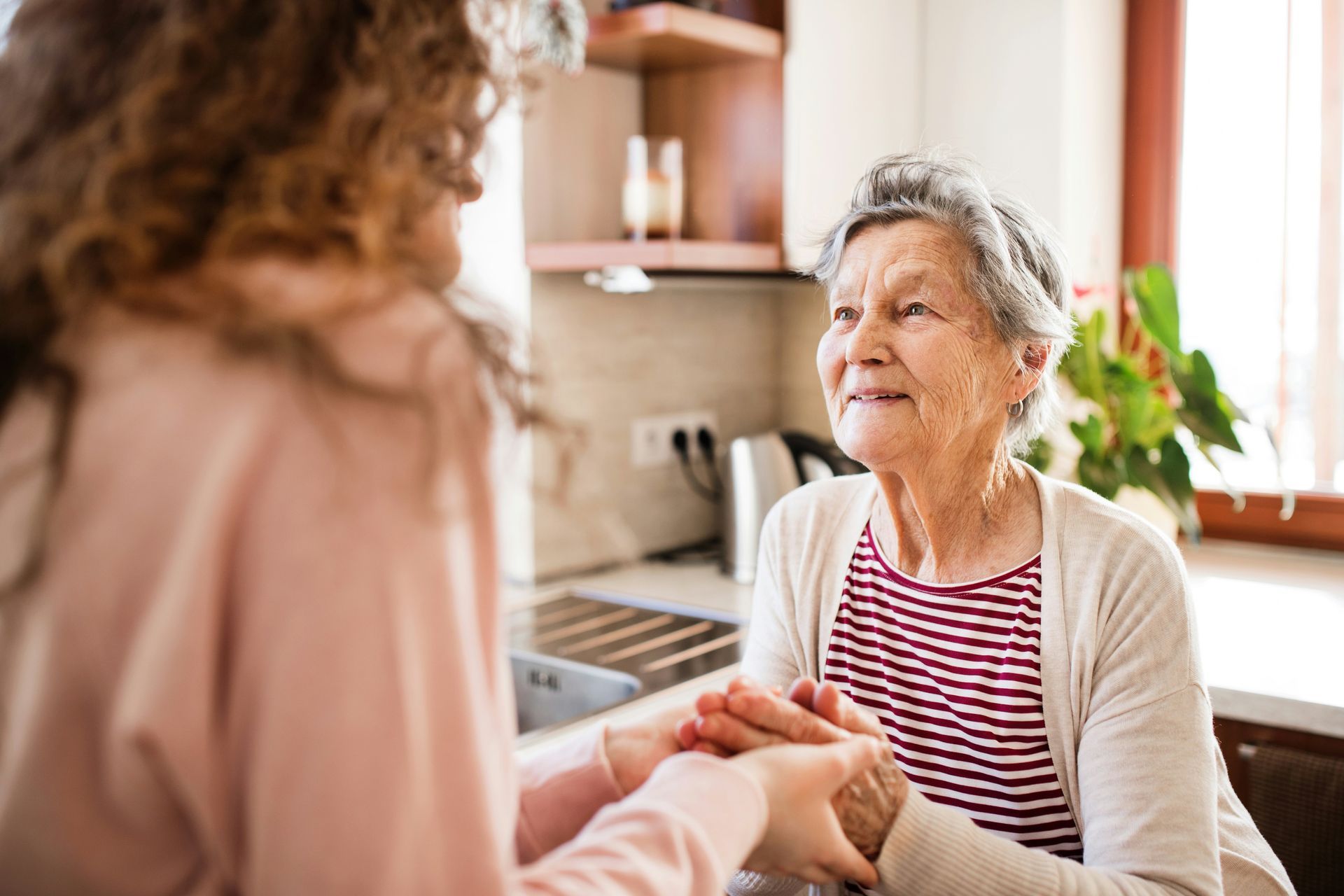 Caregiver holding an elderly woman's hands in a bright kitchen, both facing each other warmly.