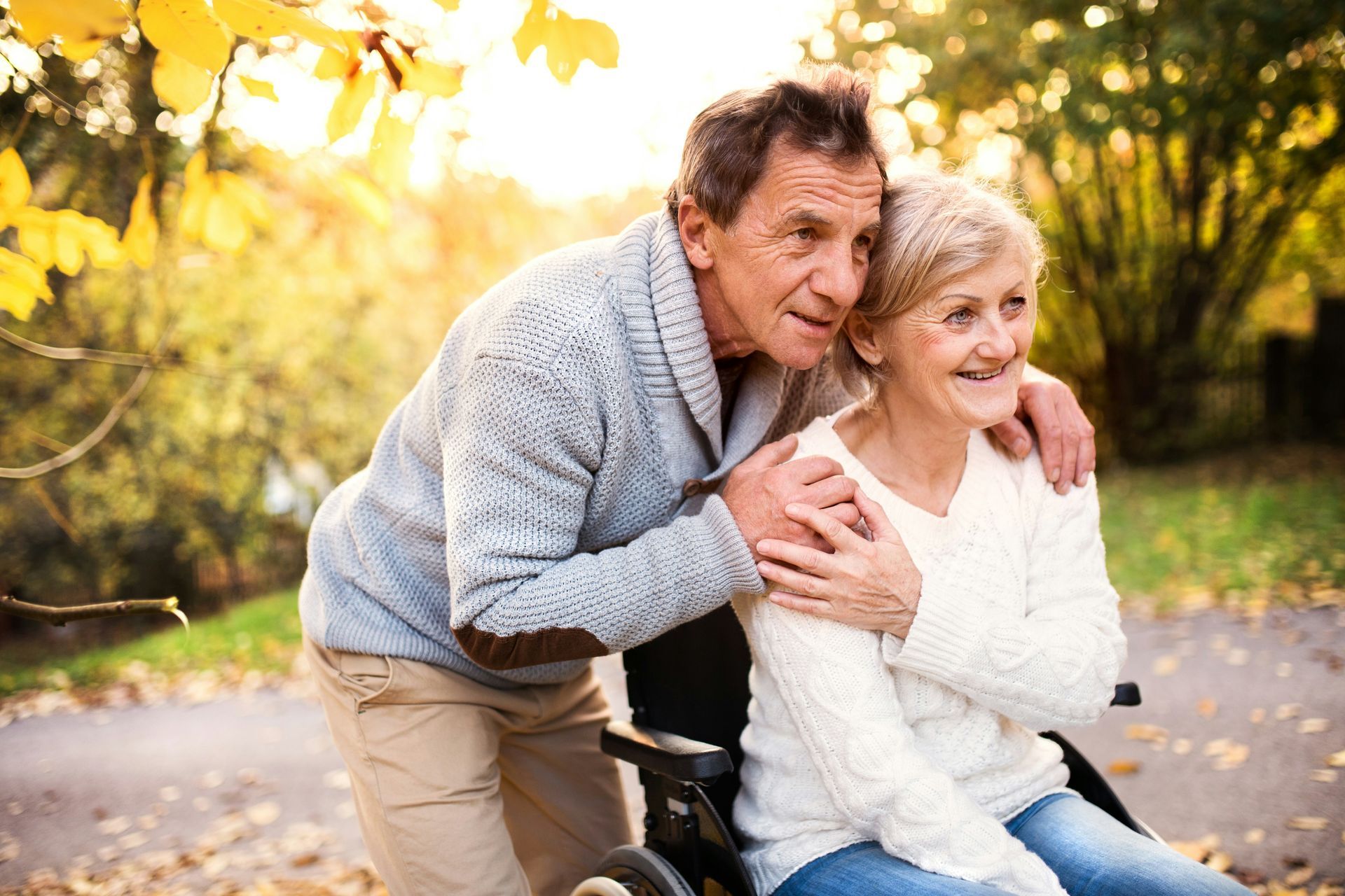 Two people smiling on a bench in an autumn park, with one arm around the other.