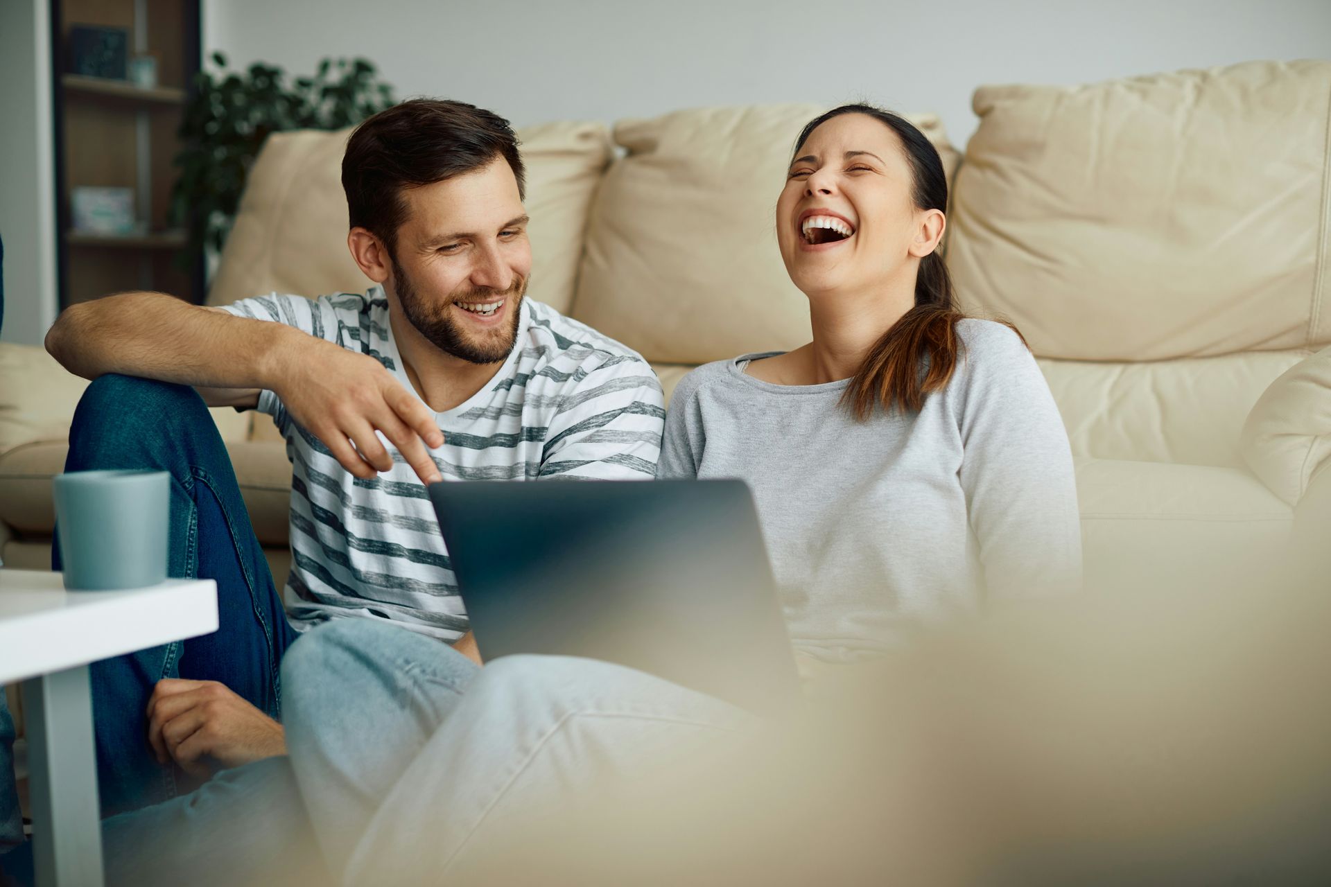 Two people laughing on a couch with a laptop in a cozy living room